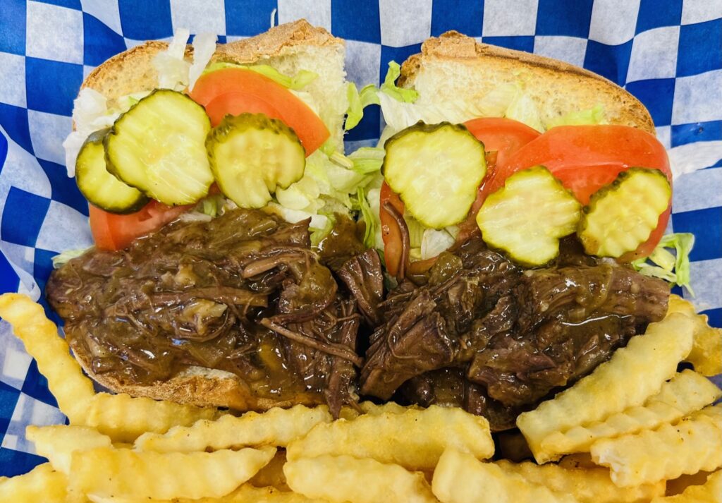Close up of shredded beef in gravy with pickles, tomatoes, and lettuce on a fresh sub roll with crinkle cut french fries