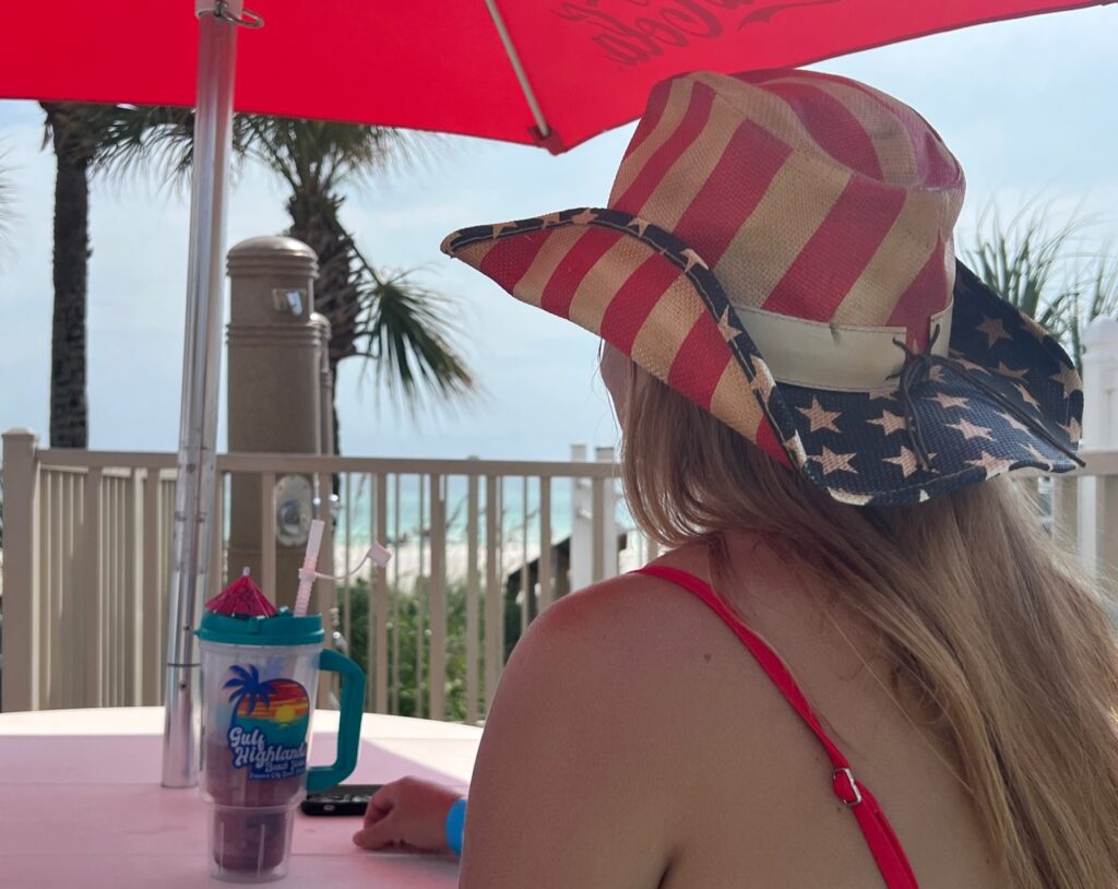Woman wearing a red, white, and blue cowboy hat enjoying a drink under a red umbrella on a patio overlooking the beach