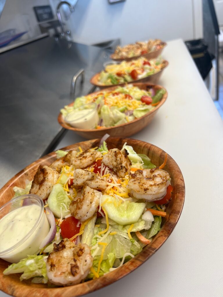 A row of grilled shrimp salads with cucumbers, carrots, and tomatoes with a side of dressing